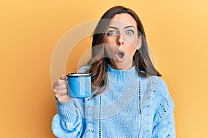 Young brunette woman holding a cup of coffee scared and amazed with open mouth for surprise, disbelief face