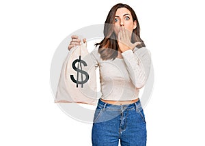 Young brunette woman holding dollars bag covering mouth with hand, shocked and afraid for mistake