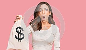 Young brunette woman holding dollars bag scared and amazed with open mouth for surprise, disbelief face