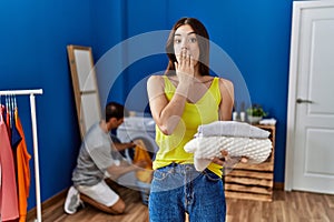 Young brunette woman holding folded laundry covering mouth with hand, shocked and afraid for mistake