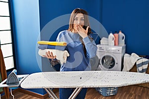 Young brunette woman holding folded laundry after ironing covering mouth with hand, shocked and afraid for mistake