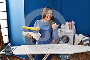 Young brunette woman holding folded laundry after ironing pointing thumb up to the side smiling happy with open mouth