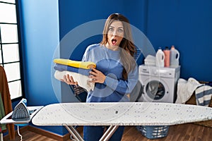 Young brunette woman holding folded laundry after ironing scared and amazed with open mouth for surprise, disbelief face