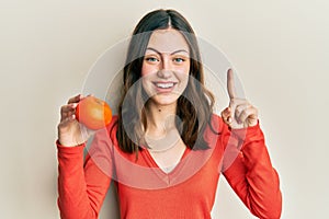 Young brunette woman holding fresh orange smiling with an idea or question pointing finger with happy face, number one