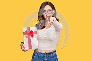 Young brunette woman holding gift pointing with finger to the camera and to you, confident gesture looking serious