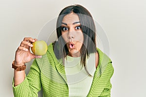 Young brunette woman holding green apple scared and amazed with open mouth for surprise, disbelief face