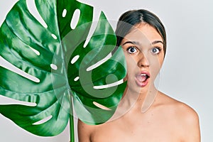 Young brunette woman holding green plant leaf close to beautiful face afraid and shocked with surprise and amazed expression, fear