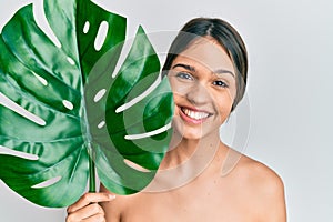 Young brunette woman holding green plant leaf close to beautiful face smiling with a happy and cool smile on face