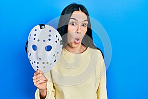 Young brunette woman holding hockey mask scared and amazed with open mouth for surprise, disbelief face