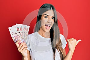 Young brunette woman holding hong kong 100 dollars banknotes pointing thumb up to the side smiling happy with open mouth