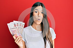 Young brunette woman holding hong kong 100 dollars banknotes scared and amazed with open mouth for surprise, disbelief face