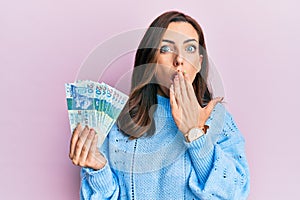 Young brunette woman holding hong kong 20 dollars banknotes covering mouth with hand, shocked and afraid for mistake