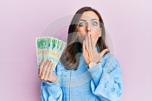 Young brunette woman holding hong kong 50 dollars banknotes covering mouth with hand, shocked and afraid for mistake