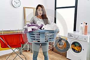Young brunette woman holding laundry basket afraid and shocked with surprise and amazed expression, fear and excited face