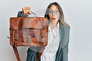 Young brunette woman holding leather bag scared and amazed with open mouth for surprise, disbelief face