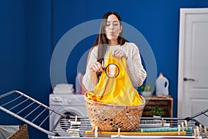 Young brunette woman holding magnifying glass looking for stain at clothes making fish face with mouth and squinting eyes, crazy