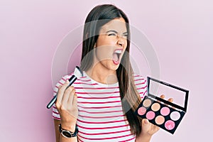Young brunette woman holding makeup brush and blush palette angry and mad screaming frustrated and furious, shouting with anger