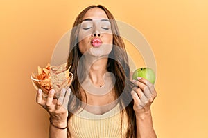 Young brunette woman holding nachos and healthy green apple looking at the camera blowing a kiss being lovely and sexy