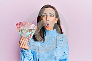 Young brunette woman holding 100 new zealand dollars banknote scared and amazed with open mouth for surprise, disbelief face