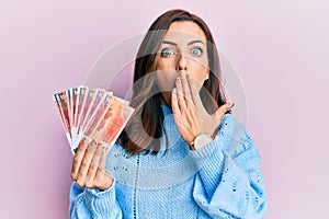 Young brunette woman holding norwegian krone banknotes covering mouth with hand, shocked and afraid for mistake