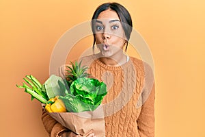 Young brunette woman holding paper bag with bread and groceries scared and amazed with open mouth for surprise, disbelief face