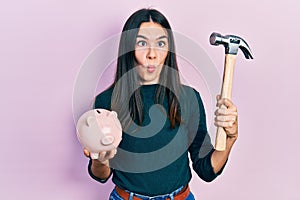 Young brunette woman holding piggy bank and hammer making fish face with mouth and squinting eyes, crazy and comical