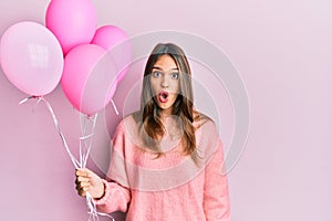 Young brunette woman holding pink balloons scared and amazed with open mouth for surprise, disbelief face