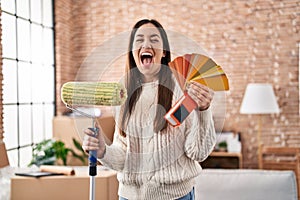 Young brunette woman holding roller painter and paint samples celebrating crazy and amazed for success with open eyes screaming