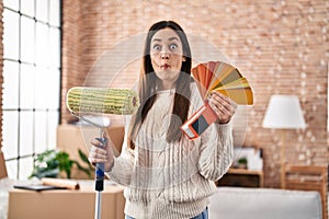 Young brunette woman holding roller painter and paint samples making fish face with mouth and squinting eyes, crazy and comical