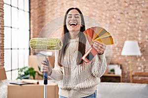 Young brunette woman holding roller painter and paint samples smiling and laughing hard out loud because funny crazy joke