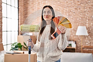 Young brunette woman holding roller painter and paint samples smiling looking to the side and staring away thinking