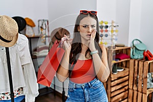 Young brunette woman holding shopping bags at retail shop afraid and shocked, surprise and amazed expression with hands on face