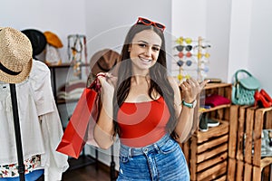 Young brunette woman holding shopping bags at retail shop pointing to the back behind with hand and thumbs up, smiling confident