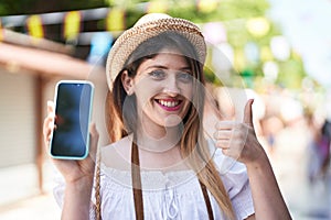 Young brunette woman holding smartphone showing blank screen smiling happy and positive, thumb up doing excellent and approval