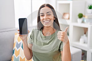 Young brunette woman holding smartphone showing blank screen smiling happy and positive, thumb up doing excellent and approval