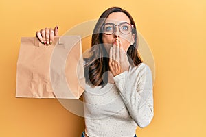Young brunette woman holding take away paper bag covering mouth with hand, shocked and afraid for mistake