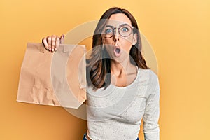 Young brunette woman holding take away paper bag scared and amazed with open mouth for surprise, disbelief face