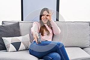 Young brunette woman holding television remote control smiling happy doing ok sign with hand on eye looking through fingers