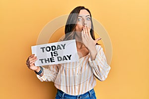 Young brunette woman holding today is the day covering mouth with hand, shocked and afraid for mistake