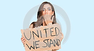 Young brunette woman holding united we stand banner covering mouth with hand, shocked and afraid for mistake
