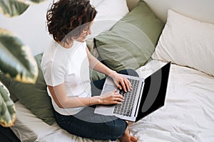 Young brunette woman in home clothes working on laptop on bed