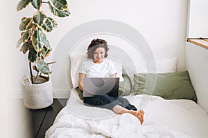 Young brunette woman in home clothes working on laptop on bed