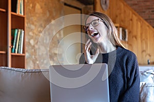 Young brunette woman laughing and talking on the smartphone in front of the laptop sitting on the sofa at home