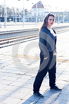 Young brunette woman in leather jacket waiting on platform at train station