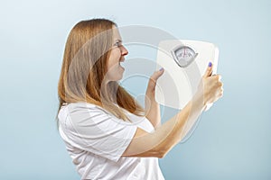 Young brunette woman looking at weight scale in shock on blue background