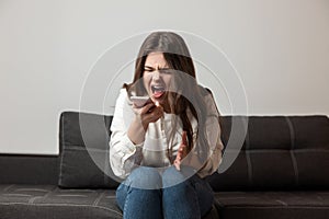 Young brunette woman looks angry hysterically shouting loud while sitting near sofa in her appartment and having