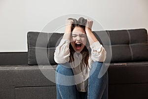 Young brunette woman looks exhausted hysterically shouting loud while sitting near sofa in her appartment, emotional