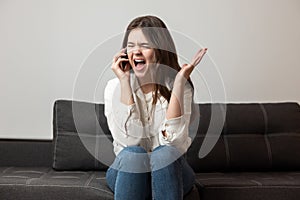 Young brunette woman looks exhausted hysterically shouting loud while sitting near sofa in her appartment and having