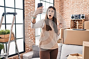 Young brunette woman moving to a new home doing video call in shock face, looking skeptical and sarcastic, surprised with open