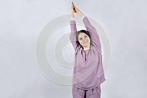Young brunette woman in muslin lavender pajamas sleep clothes stretches by raising her arms up.